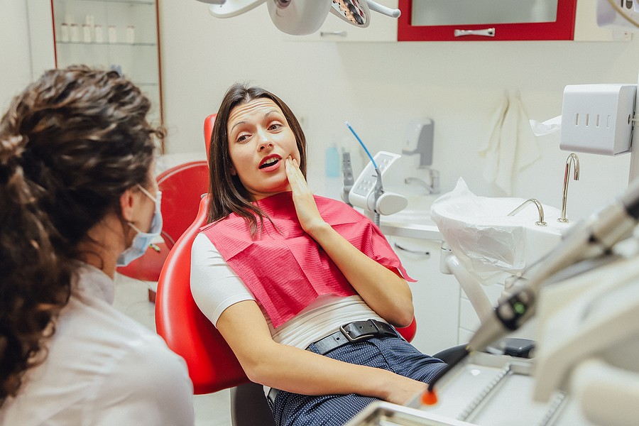A woman sitting in a dental chair holding her jaw in pain while speaking with a dentist about a tooth abscess.