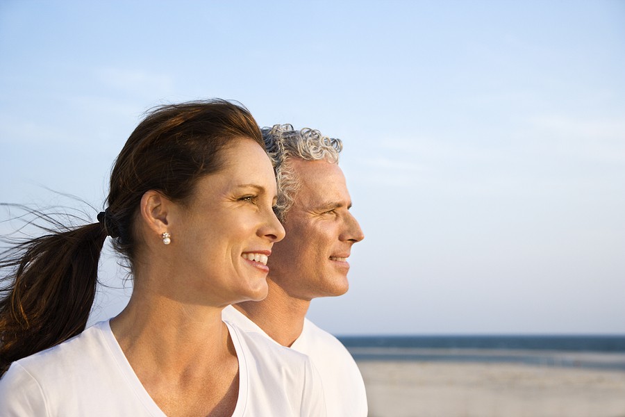 Smiling middle-aged couple enjoying the beach, looking toward the ocean under a clear sky.